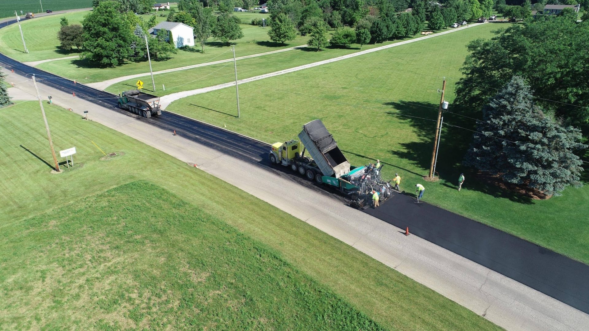 Aerial view of a road being paved by construction workers and heavy machinery, with green lawns, trees, and a white house in the background on a sunny day. Aerial view of a road being paved by construction workers and heavy machinery, with green lawns, trees, and a white house in the background on a sunny day.