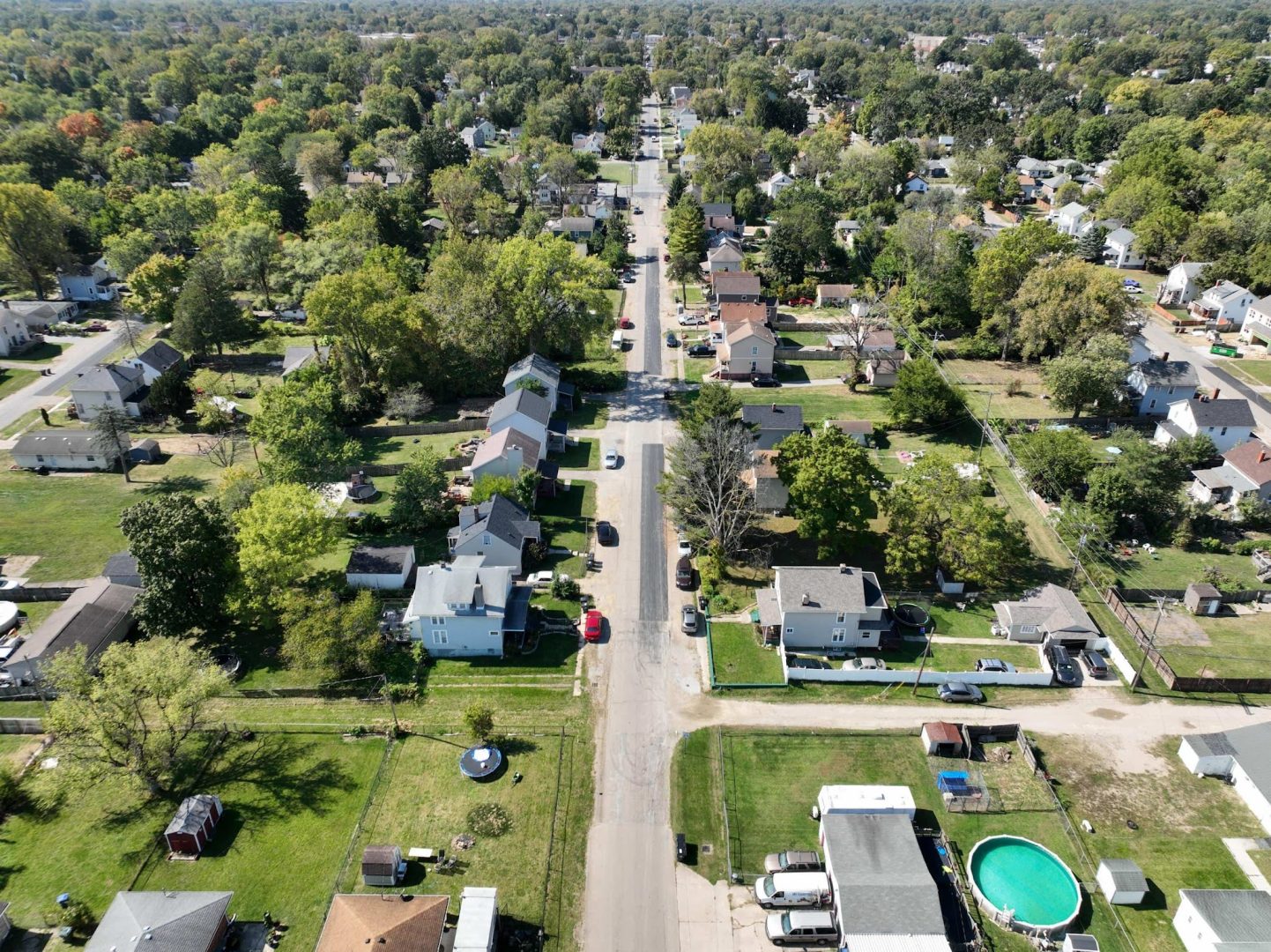 Aerial view of a residential neighborhood with houses, trees, lawns, and a few cars parked along a straight street. There is a round above-ground pool in one backyard and the area is surrounded by greenery. Aerial view of a residential neighborhood with houses, trees, lawns, and a few cars parked along a straight street. There is a round above-ground pool in one backyard and the area is surrounded by greenery.