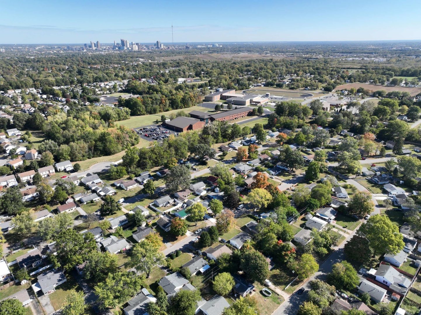 Aerial view of a suburban neighborhood with tree-lined streets and single-family homes, a large school complex in the center, and a distant city skyline under a clear blue sky. Aerial view of a suburban neighborhood with tree-lined streets and single-family homes, a large school complex in the center, and a distant city skyline under a clear blue sky.