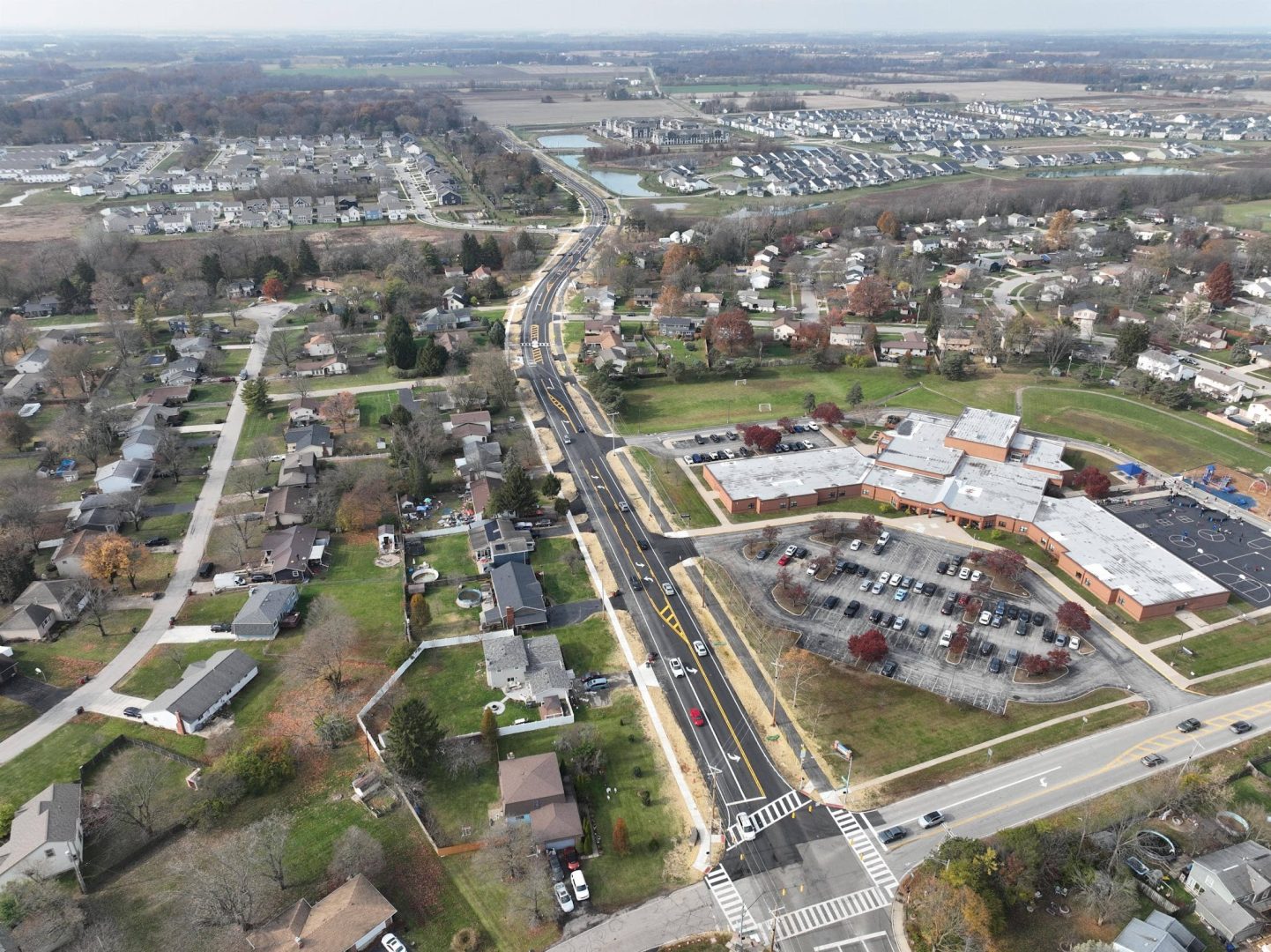 Aerial view of a suburban neighborhood with houses, a school with a parking lot, streets with vehicles, and open fields in the background. Trees with autumn foliage are scattered throughout the area.