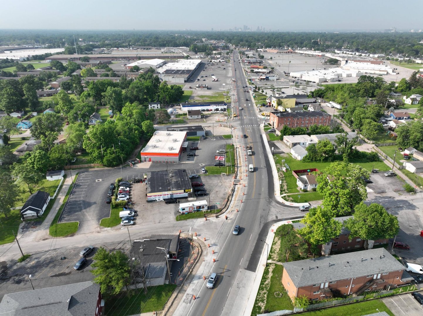 Aerial view of a busy urban street lined with businesses, stores, parking lots, and residential buildings, with trees scattered throughout and cars driving along the road under a hazy sky. Aerial view of a busy urban street lined with businesses, stores, parking lots, and residential buildings, with trees scattered throughout and cars driving along the road under a hazy sky.