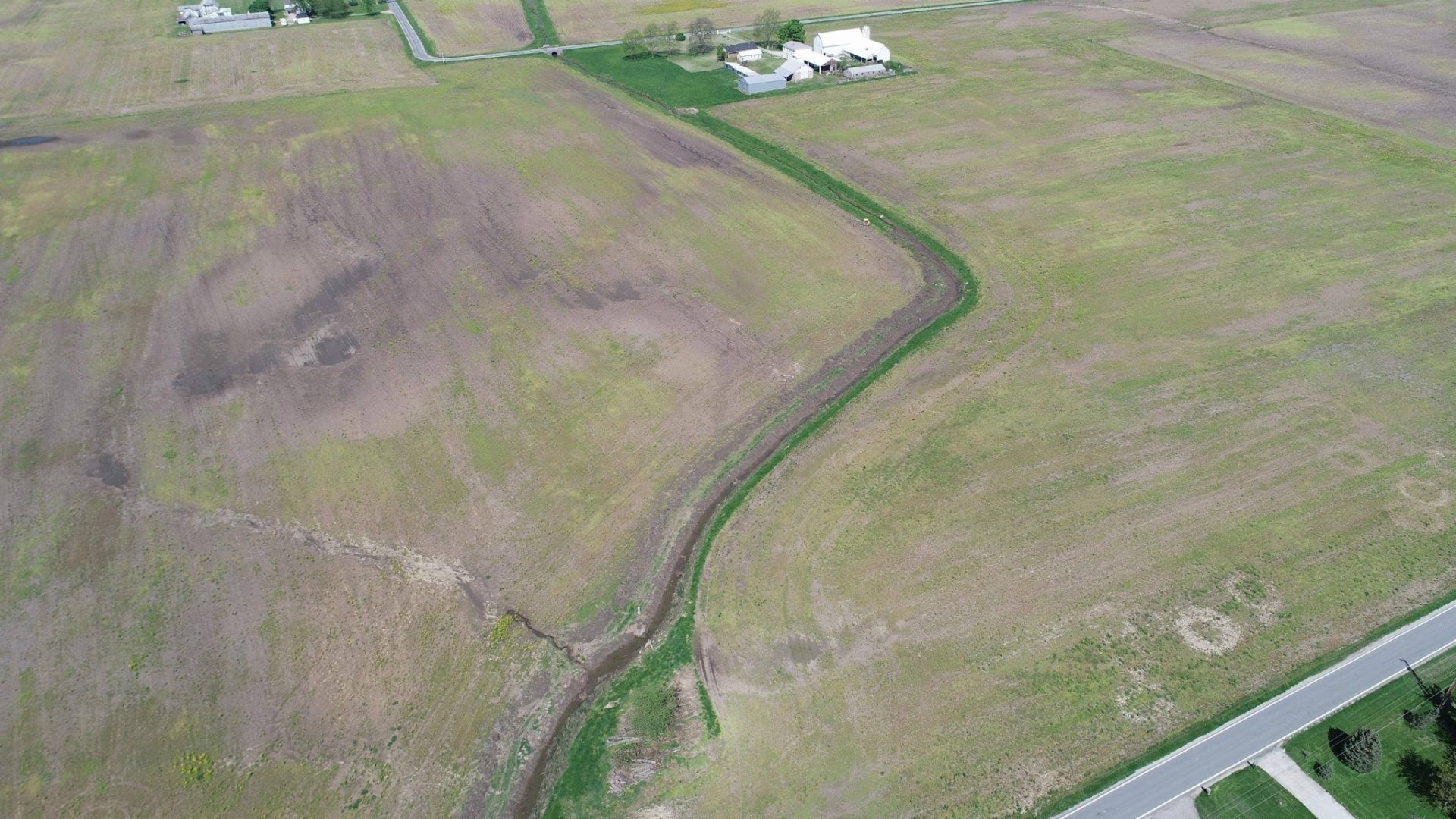 Aerial view of a large, mostly empty green field with a winding dirt path or stream running through it, a few buildings near the top, and a road along the bottom right edge. Aerial view of a large, mostly empty green field with a winding dirt path or stream running through it, a few buildings near the top, and a road along the bottom right edge.