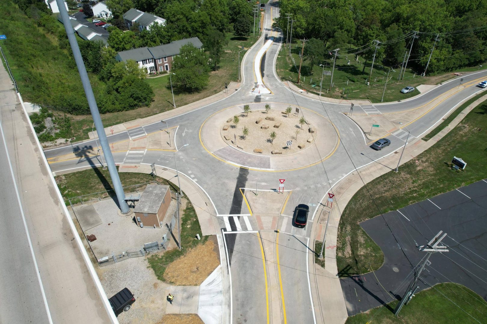 Aerial view of a roundabout intersection with several cars navigating, crosswalks, landscaped center, nearby buildings, trees, and a parking lot. Multiple roads connect to the roundabout. Aerial view of a roundabout intersection with several cars navigating, crosswalks, landscaped center, nearby buildings, trees, and a parking lot. Multiple roads connect to the roundabout.