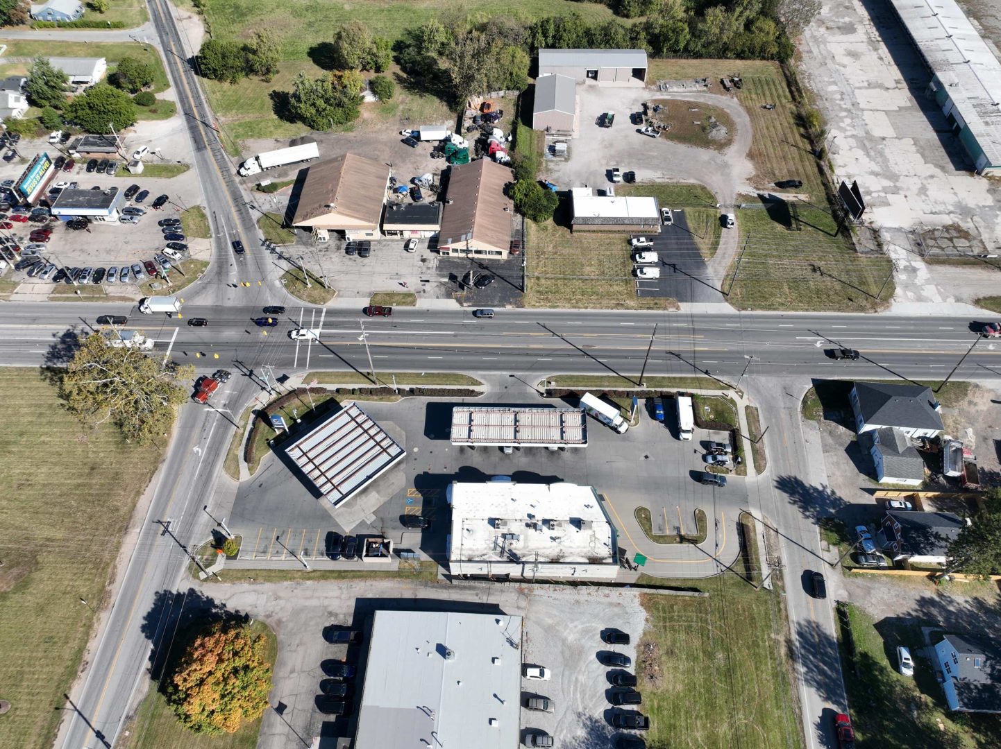 Aerial view of a commercial area with multiple buildings, parking lots, cars, and a main road running horizontally through the center, surrounded by grassy areas and some trees. Aerial view of a commercial area with multiple buildings, parking lots, cars, and a main road running horizontally through the center, surrounded by grassy areas and some trees.