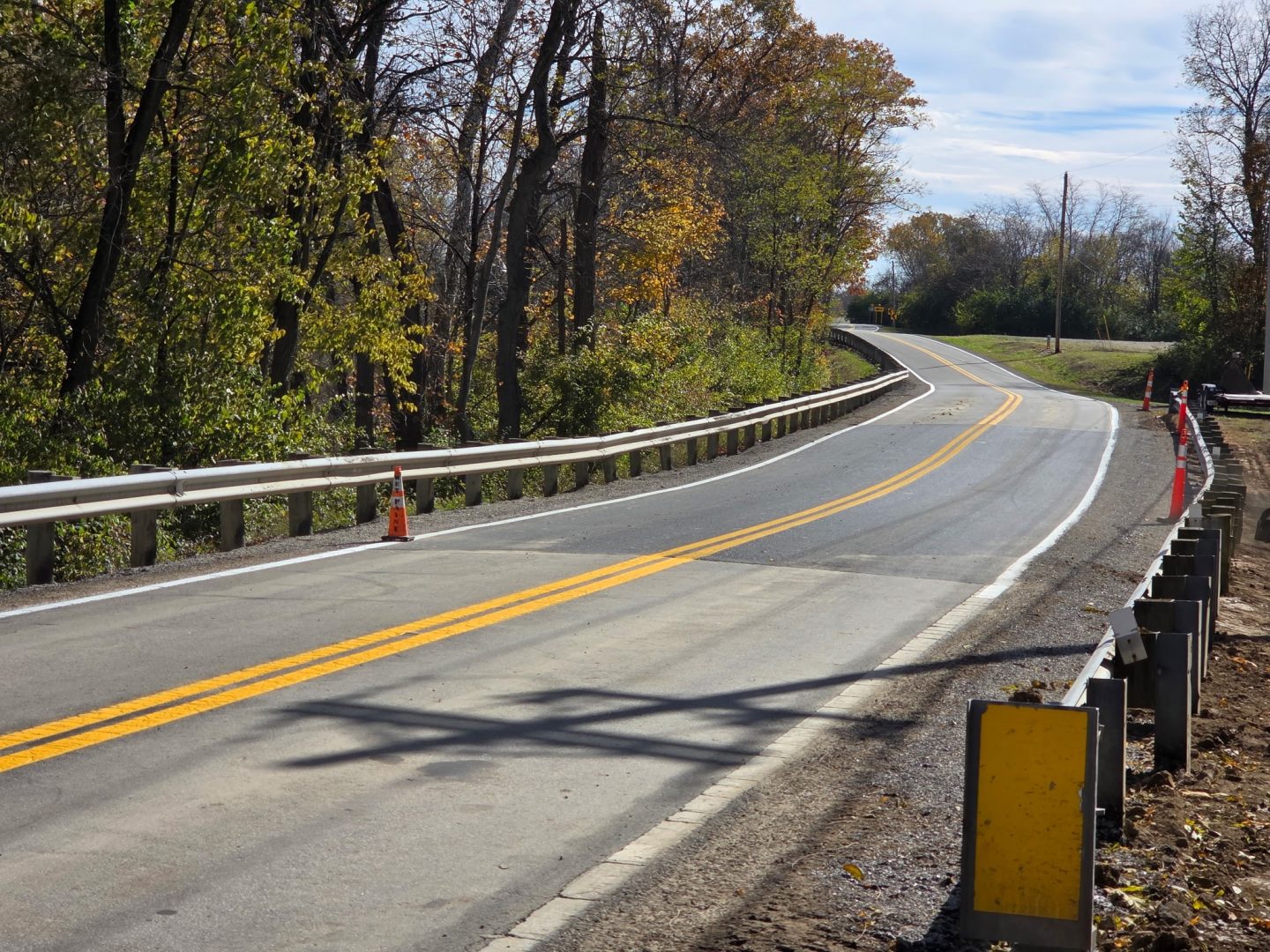A two-lane road with yellow lines curves through a wooded area. Guardrails run along both sides, and an orange traffic cone marks the shoulder. Trees with autumn leaves line the road under a clear sky. A two-lane road with yellow lines curves through a wooded area. Guardrails run along both sides, and an orange traffic cone marks the shoulder. Trees with autumn leaves line the road under a clear sky.