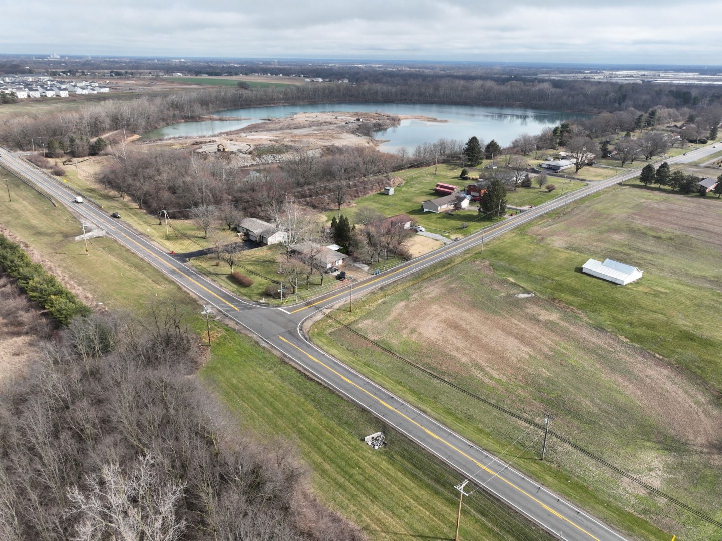 Aerial view of a rural crossroads with intersecting roads, houses, and farmland. There is a large pond or lake surrounded by trees in the background under a cloudy sky.