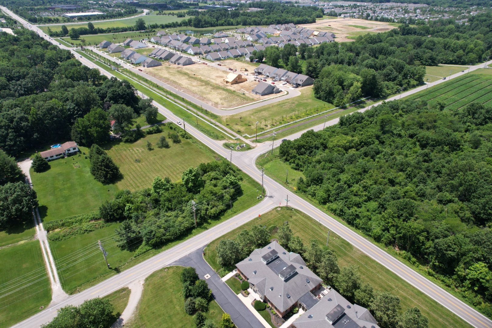 Aerial view of a suburban area with a large housing development under construction, surrounded by green trees and roads intersecting in the foreground, with homes and open fields nearby. Aerial view of a suburban area with a large housing development under construction, surrounded by green trees and roads intersecting in the foreground, with homes and open fields nearby.